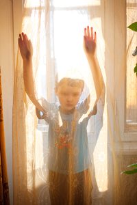 A young boy wearing a blue t-shirt and dark colored shorts stands in between a brightly lit window and its sheer curtain. With his back to the window, he looks through the curtain, lifting his arms above his head so his palms push the curtain away from him, slightly.