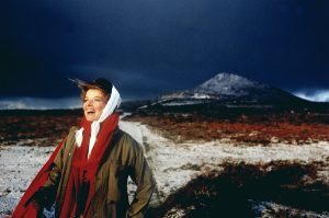 Katherine Hepburn stands in front of a mountain landscape, looking gleefully into the distance off to the left side of the image. She is wearing a green flannel shirt, long red scarf around her neck, and has a white scarf wrapped around her head.