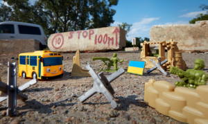 A plastic yellow school bus, plastic toy jacks, a plastic Ukrainian flag and two plastic green 'army men' solider toys are scattered on the ground. The two soldier toys aim their weapons at the bus as it approaches. In the background sits a concrete brick with the words "STOP 100m" written on it in red paint.