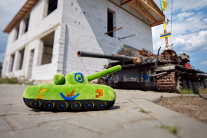 A bright green, plush toy shaped like an army tank sits in the foreground on a concrete sidewalk. In the background, a full-size rusted army tank sits outside of a white, brick building.