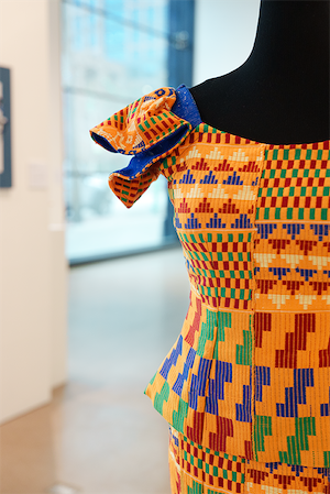 Close-up of a handwoven peplum blouse and skirt in orange, green, blue, and yellow geometric patterns with shoulder ruffles displayed on a black mannequin in a gallery space.