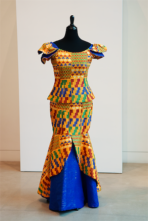 A peplum blouse and long skirt made with Kente strips from Ghana, featuring bright orange, green, blue, and yellow geometric patterns, displayed on a black mannequin with ruffled shoulders and a flared skirt revealing a blue underskirt.
