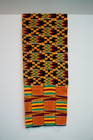 A four-paneled handwoven Kente textile from Ghana with repeating diamond, zigzag, and geometric patterns in warm black, yellow, green, and red.