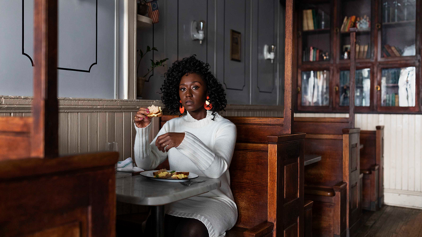 A woman in a white long sleeve dress sits in a wooden diner booth, pausing mid-sip to confront the viewer.