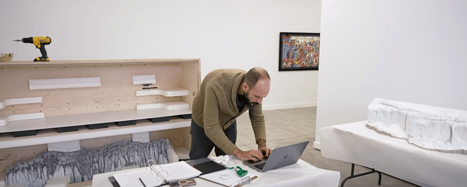 Man types on his computer in a gallery with a binder next to him and crates of artwork behind him.