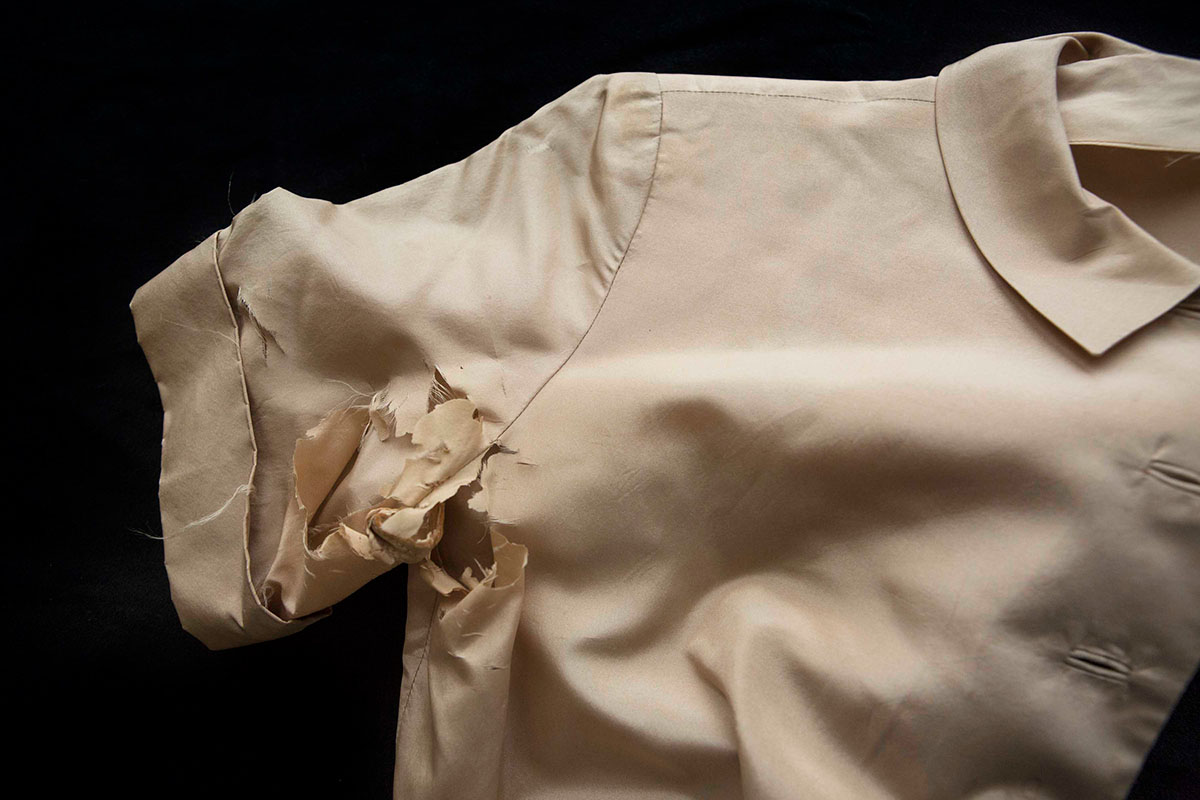 Cropped view of an off-white, wrinkled, short-sleeved collared shirt with a shredded armpit against a black background.