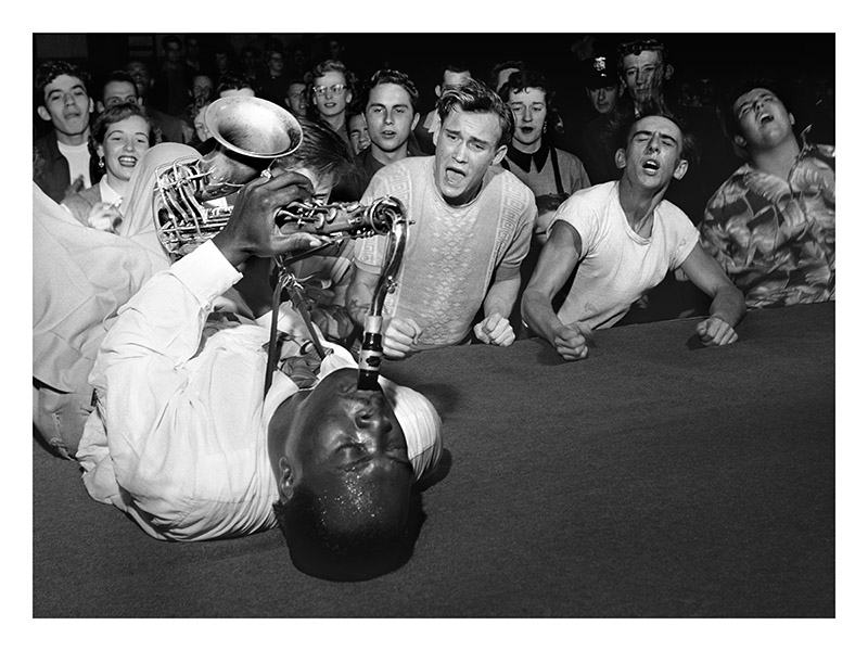 Big Jay McNeely plays a saxophone lying at the edge of a stage, the crowd pressed up against it.