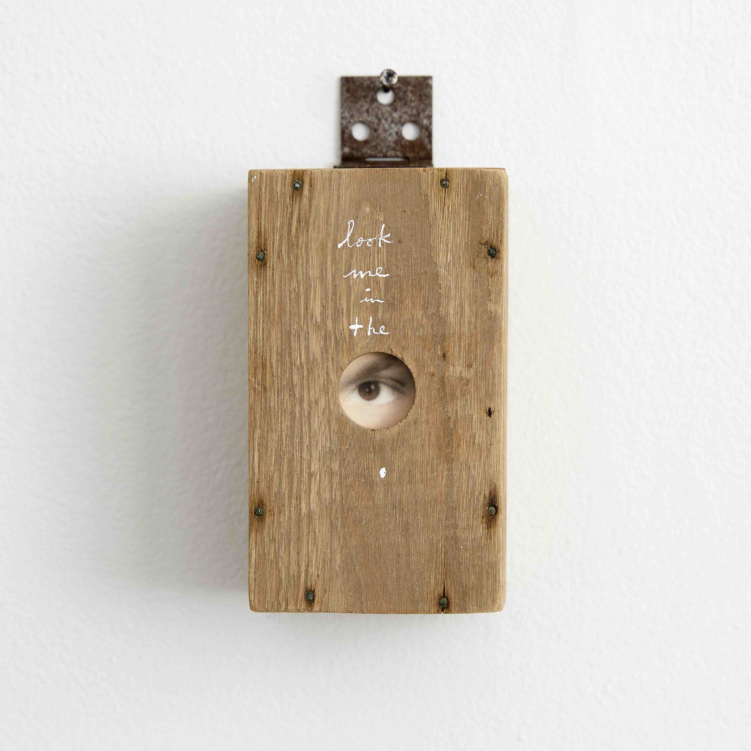 A wooden box hangs on a wall by a rusted hinge. At the center of the box, a hole is cut and a vintage photograph peeks through, exposing only a brown eye and eyebrow. Above the hole, white cursive letters read “Look me in the”.