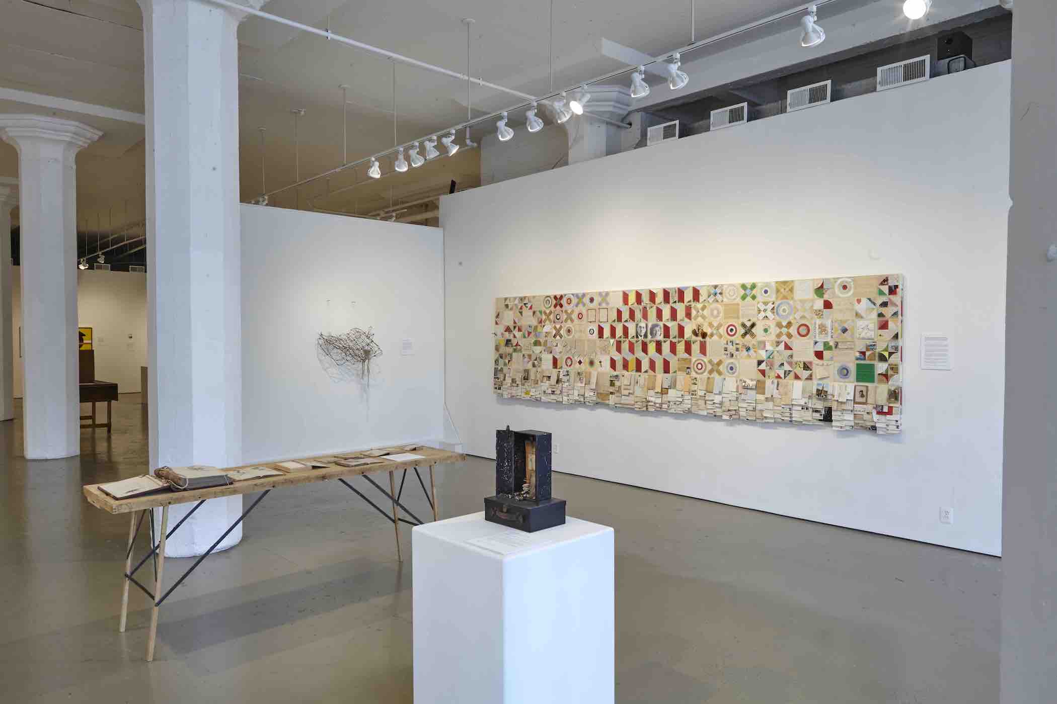 A long brown wooden table sits in the middle of an art gallery. Vintage books are laid open across the table. In the foreground, artwork sits on a white pedestal. Artworks hangs on the white gallery walls in the background.