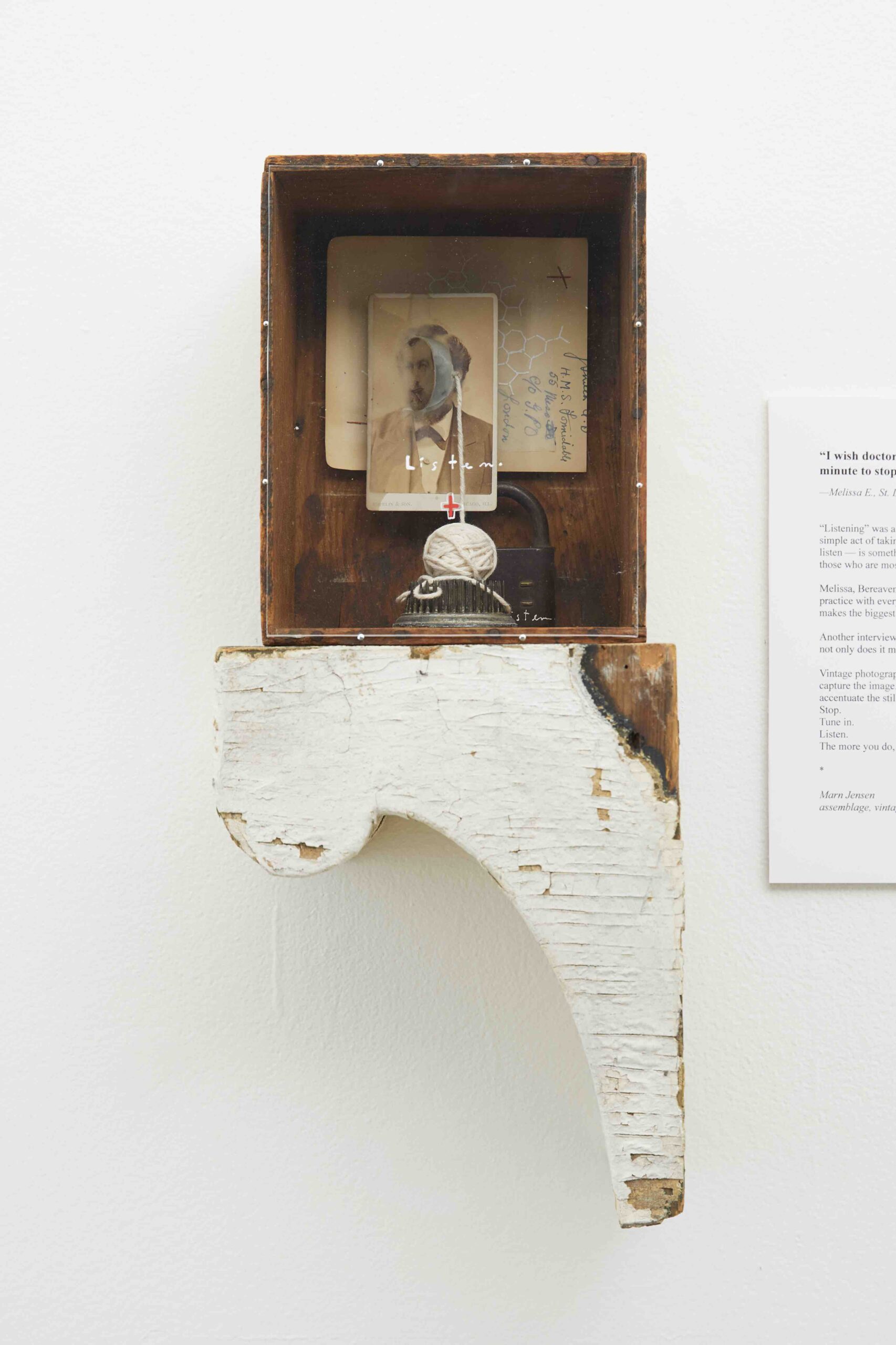 An old wooden box sits with the inside facing the viewer on a small wooden pedestal, attached to a wall. A pad lock, small ball of string, and old photograph, and other scraps of paper are displayed in the open drawer behind a piece of plexiglass. The word “Listen” is written in white letters on the plexiglass above a red cross.
