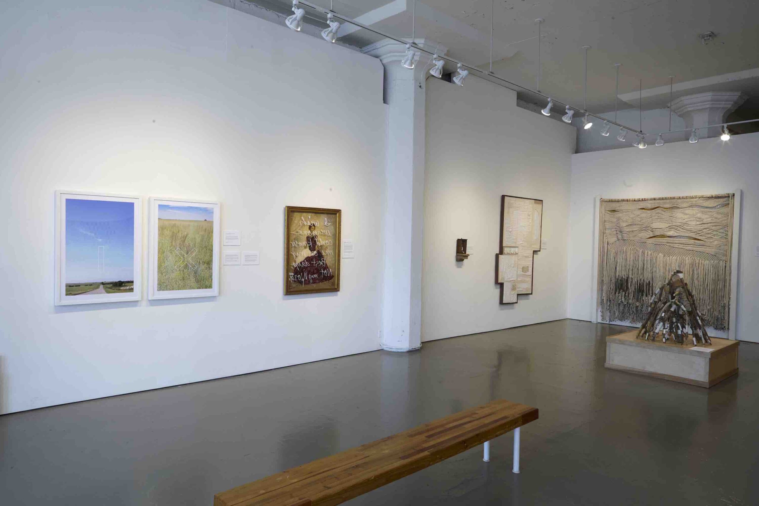 A long wooden bench sits in the middle of an art gallery. Artwork hangs on the surrounding walls and on nearby pedestals.