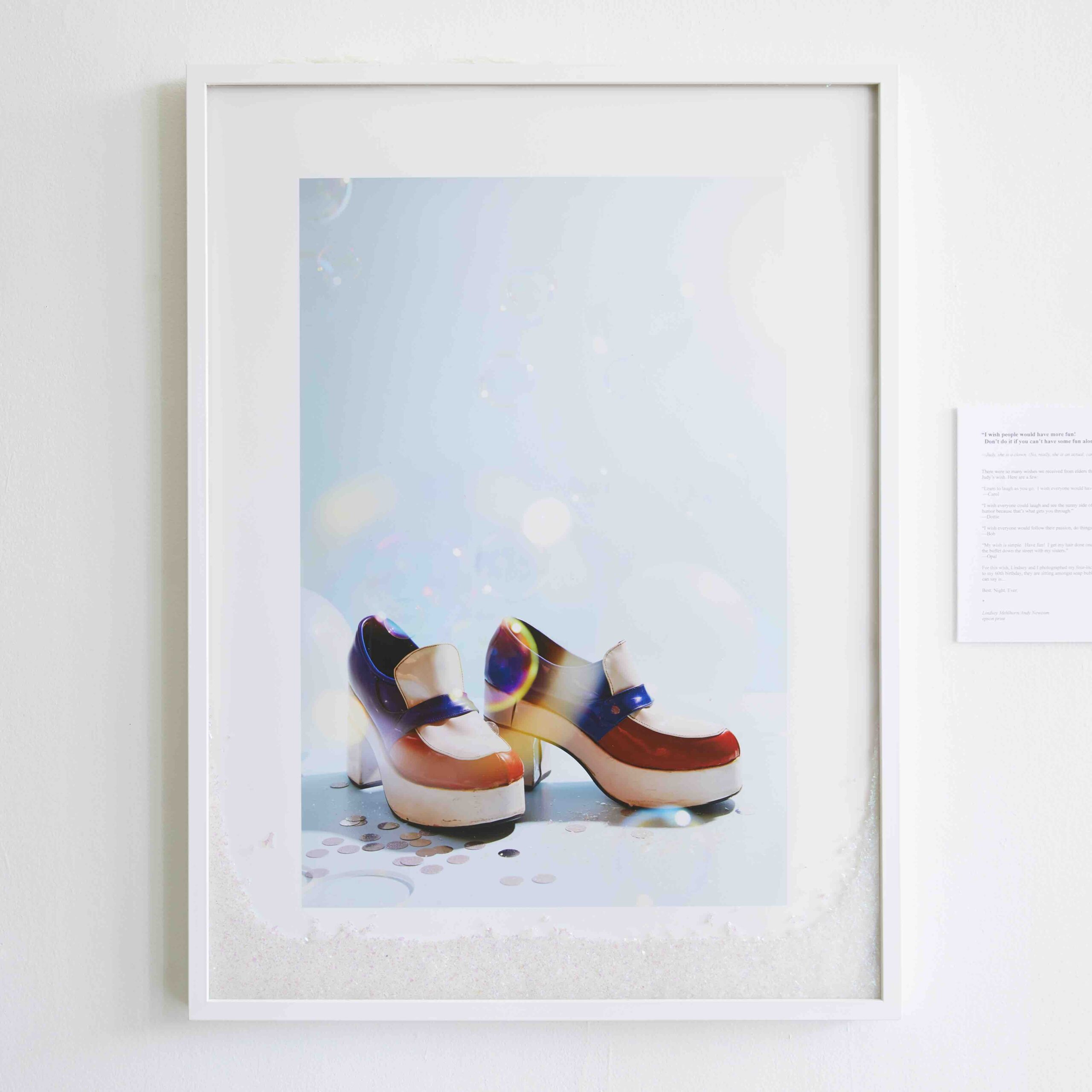 A framed photograph of multi-colored chunky heels against a blue background. On the ground near the shoes sits scattered pieces of silver confetti. Superimposed into the bottom of the frame is white glitter.