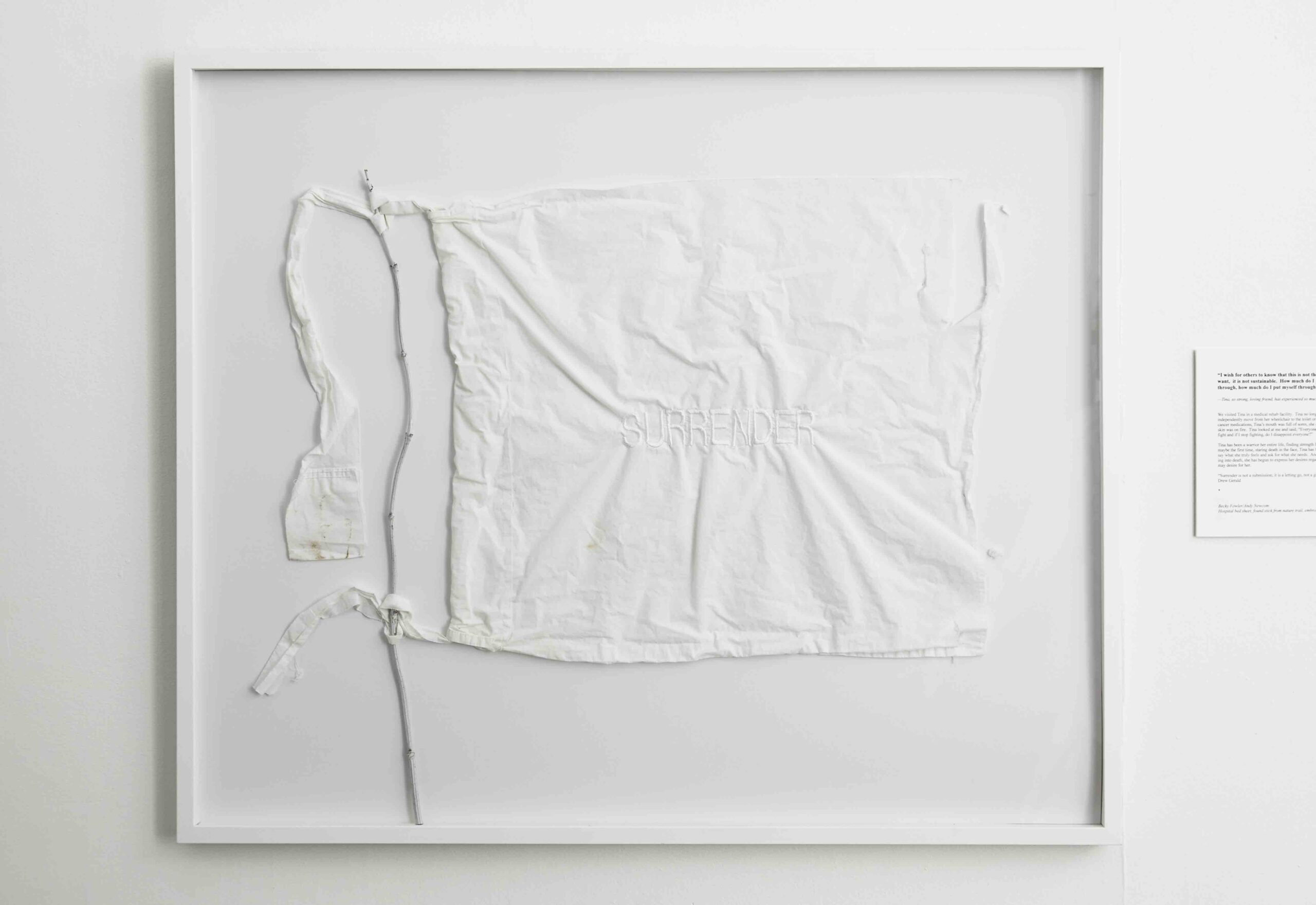 A white hospital sheet is attached to a thin white stick like a flag to a pole. The word “SURRENDER” sewn into the center of the sheet in white thread. The assemblage is framed against a white background.
