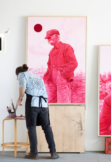 The artist, Joel Daniel Phillips, is pictured working in his art studio. Joel's back is turned towards the camera as he kneels in front of three large scale drawings--two pink oil paintings of men with nature backdrops, and another charcoal drawing depicting a young child in a cornfield.
