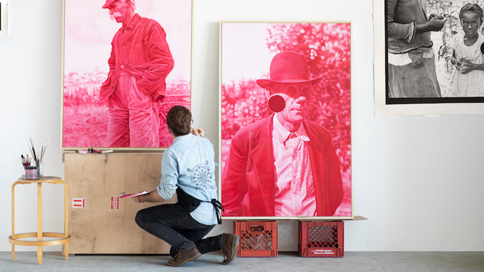 The artist, Joel Daniel Phillips, is pictured working in his art studio. Joel's back is turned towards the camera as he kneels in front of three large scale drawings--two pink oil paintings of men with nature backdrops, and another charcoal drawing depicting a young child in a cornfield.