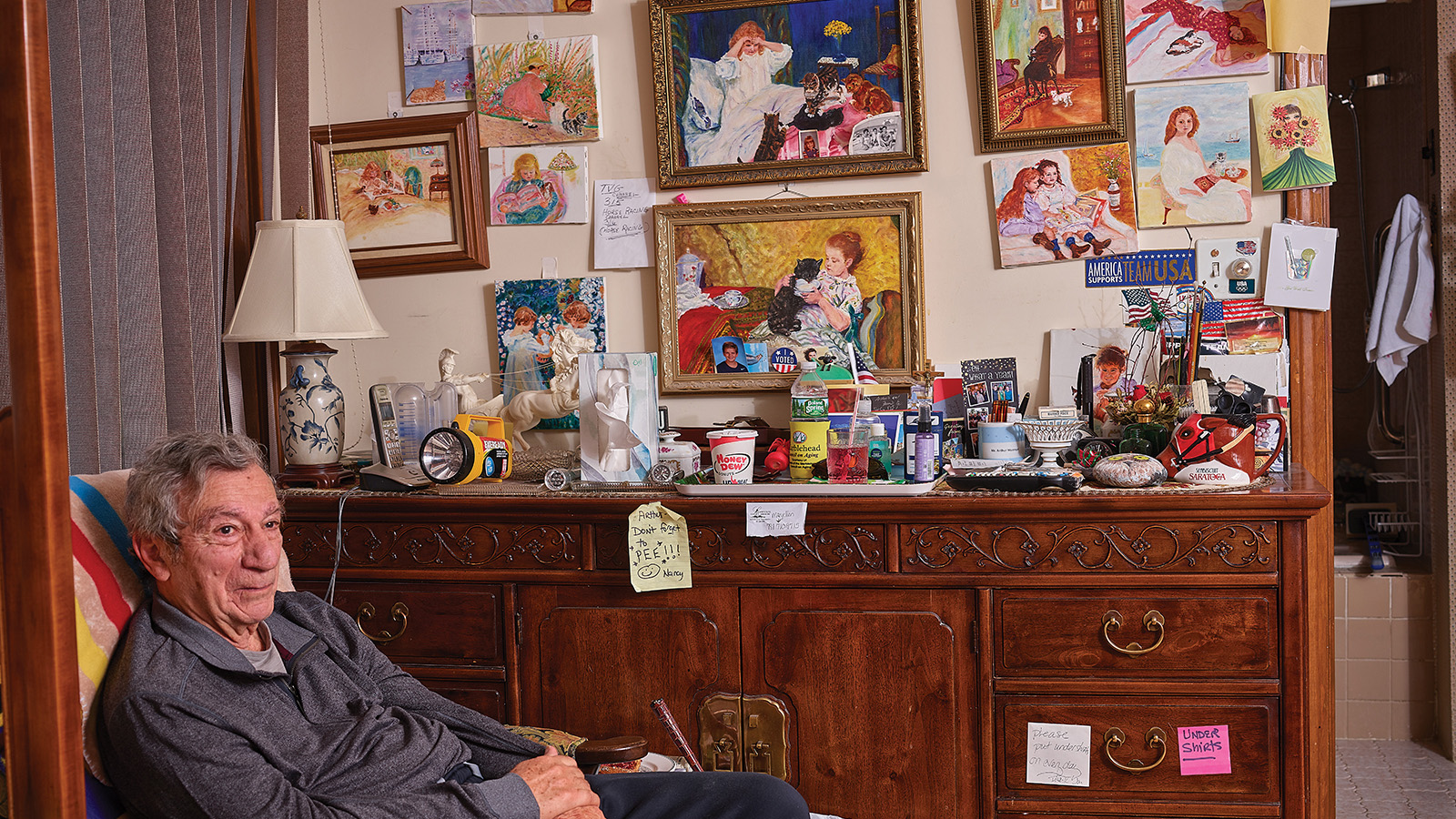 An elderly fair skinned man wearing a grey long sleeve shirt and black pants sits in a chair to the left of the photograph. On the wall behind him sits a brown wooden dresser and a wall covered in colorful artwork.