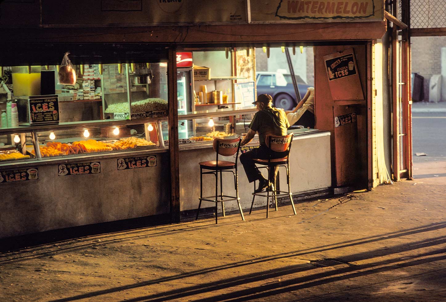 At an open-air food stand in warm evening light with long shadows, a person sits on one of two stools at the counter facing trays of food in buffet warmers.