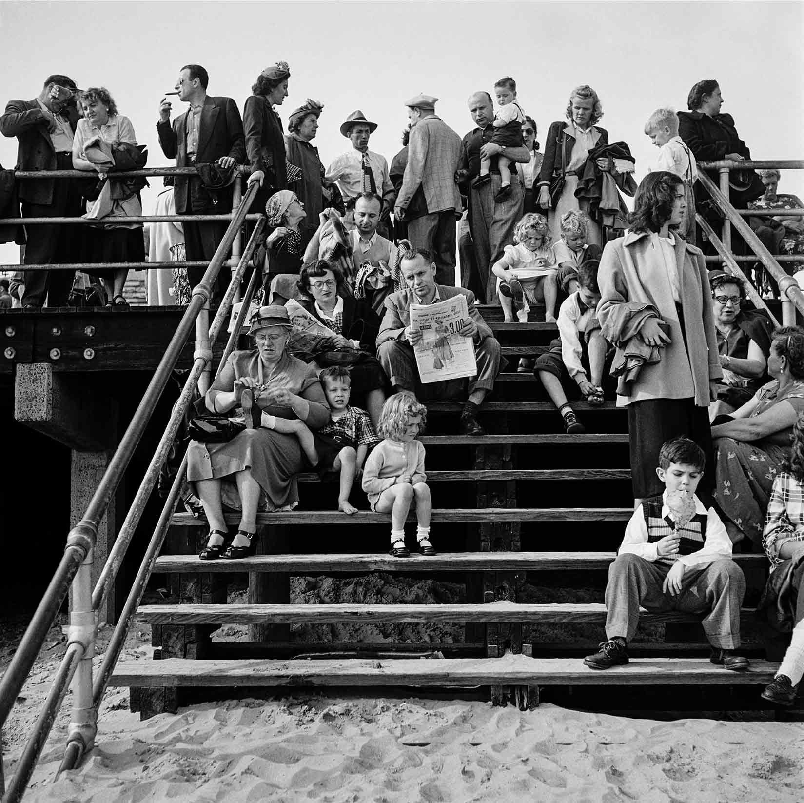 On a set of wooden steps above a sandy beach, people of varying ages spend a leisurely time chatting, reading, eating, and tending to children, creating a crowded, layered scene.