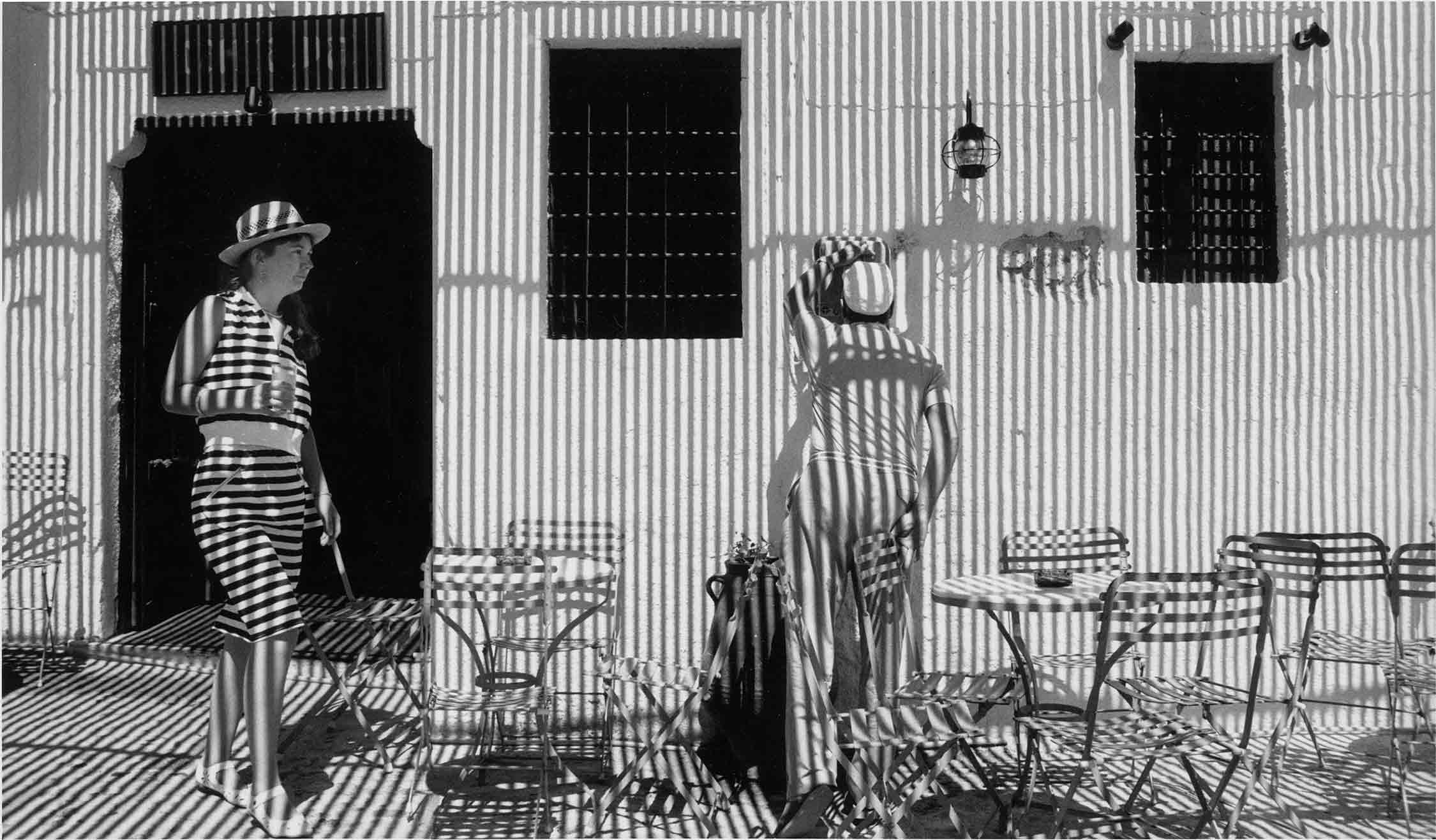 Against a white building striped with strong shadows, three women in patterned dresses move and gather among metal café chairs and tables, as the light casts bold linear patterns across their bodies and the ground.