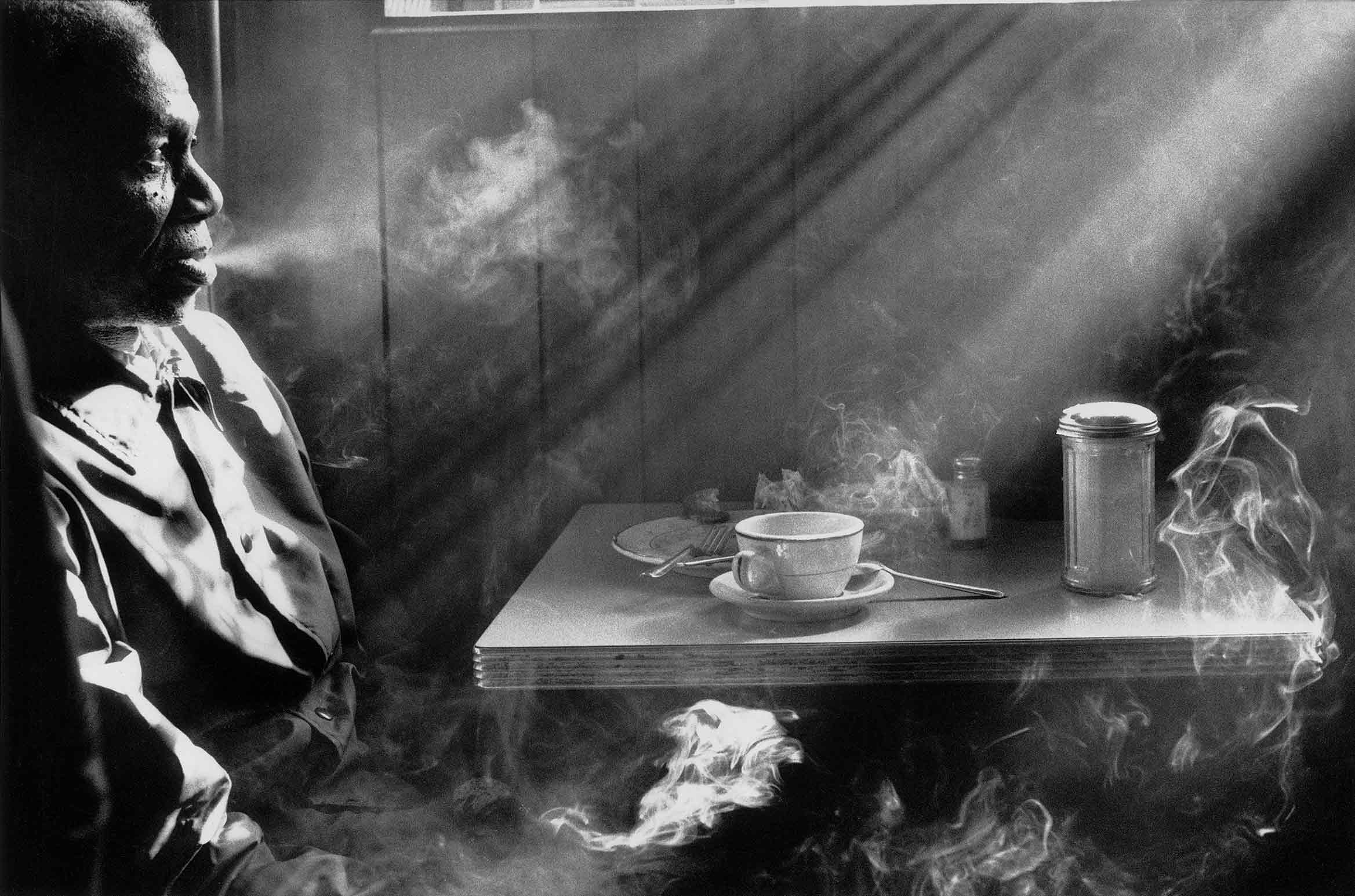 At a diner table, an older man sits across from a cup and saucer, billowing smoke from his cigarette as the sun casts bright rays of light across the scene.