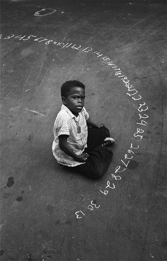 A small boy in a white shirt and black pants sits on the asphalt, pausing from drawing a long line of numbers with a piece of chalk to look up.