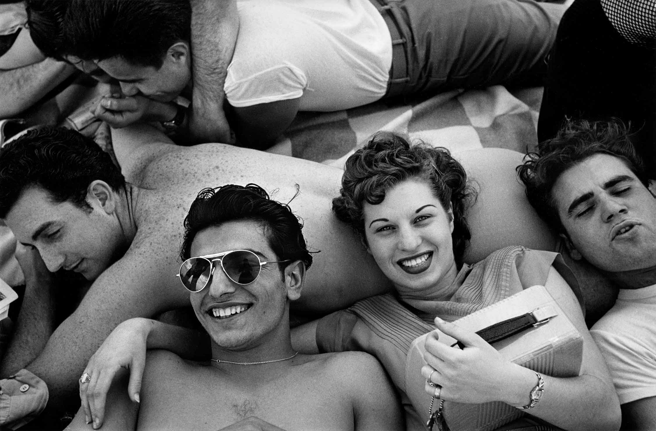 On a blanket, a small group of young people lounge closely together, some leaning on one another as they laugh and smile.