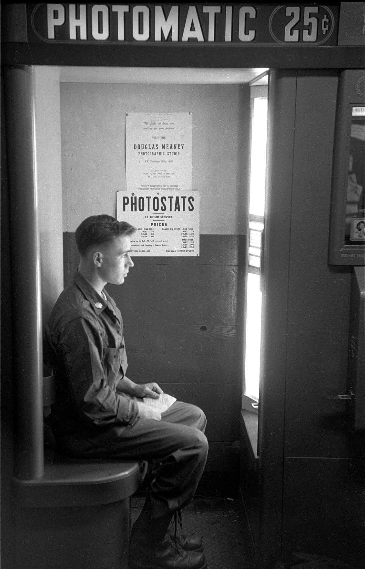 Inside a photobooth labeled ‘PHOTOMATIC 25¢,’ a young man in uniform sits in profile on a stool holding a small paper, illuminated by a bright light, with posted signs visible on the wall behind him.