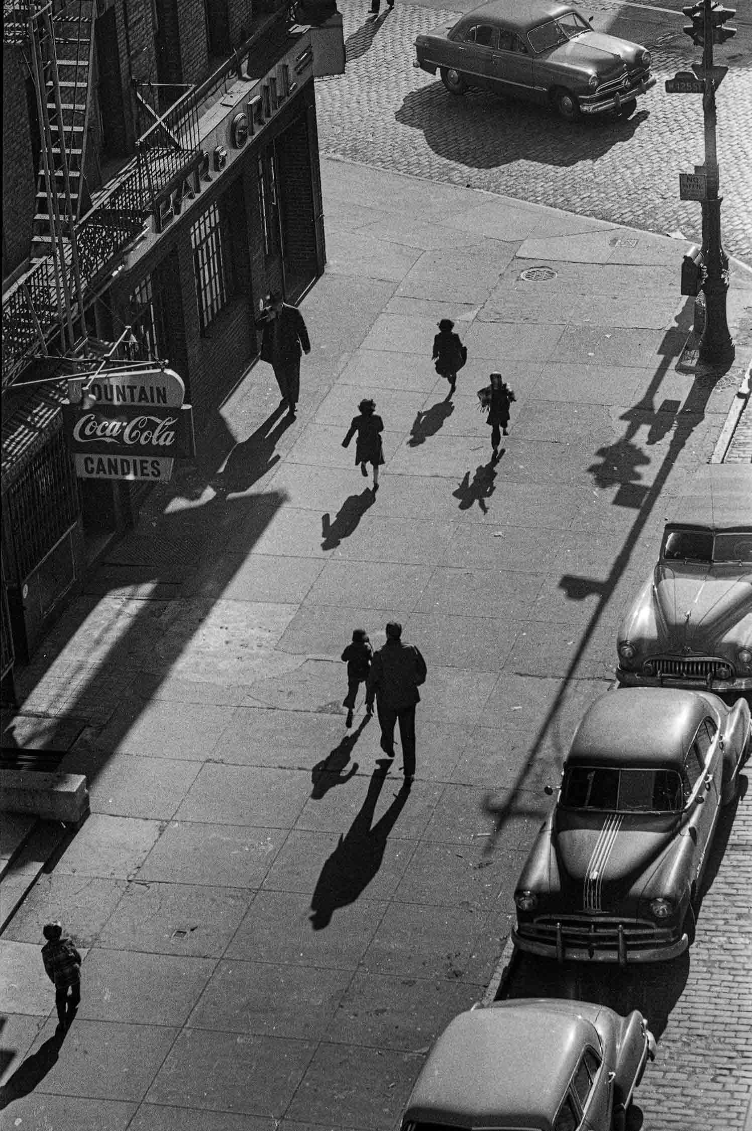 From an overhead view of a street, an adult and five children walk along a sidewalk between a Coca-Cola fountain shop and parked cars, casting long afternoon shadows.