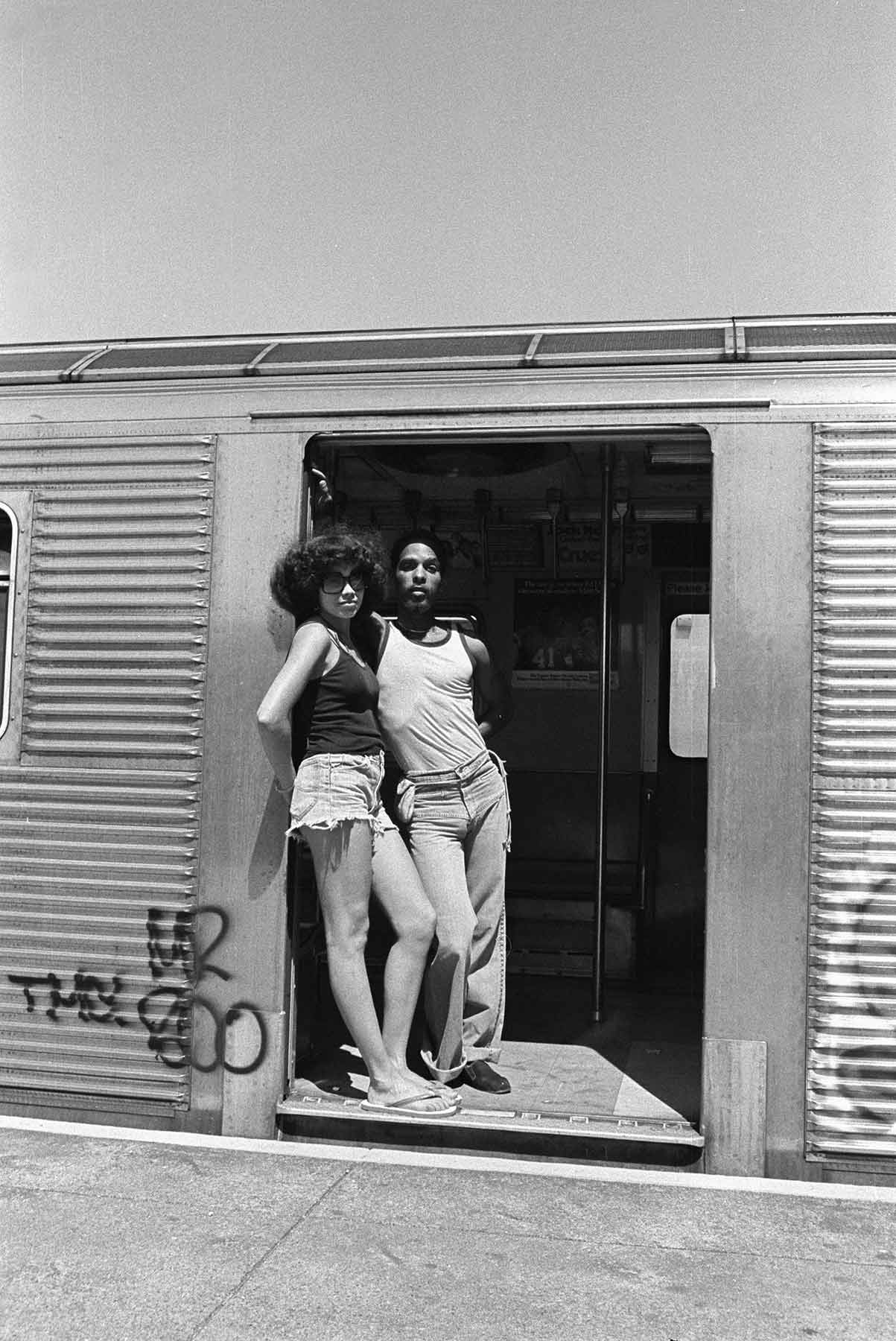 In bright afternoon sunlight, a couple dressed for summer leans close together against the doorframe of a subway car.