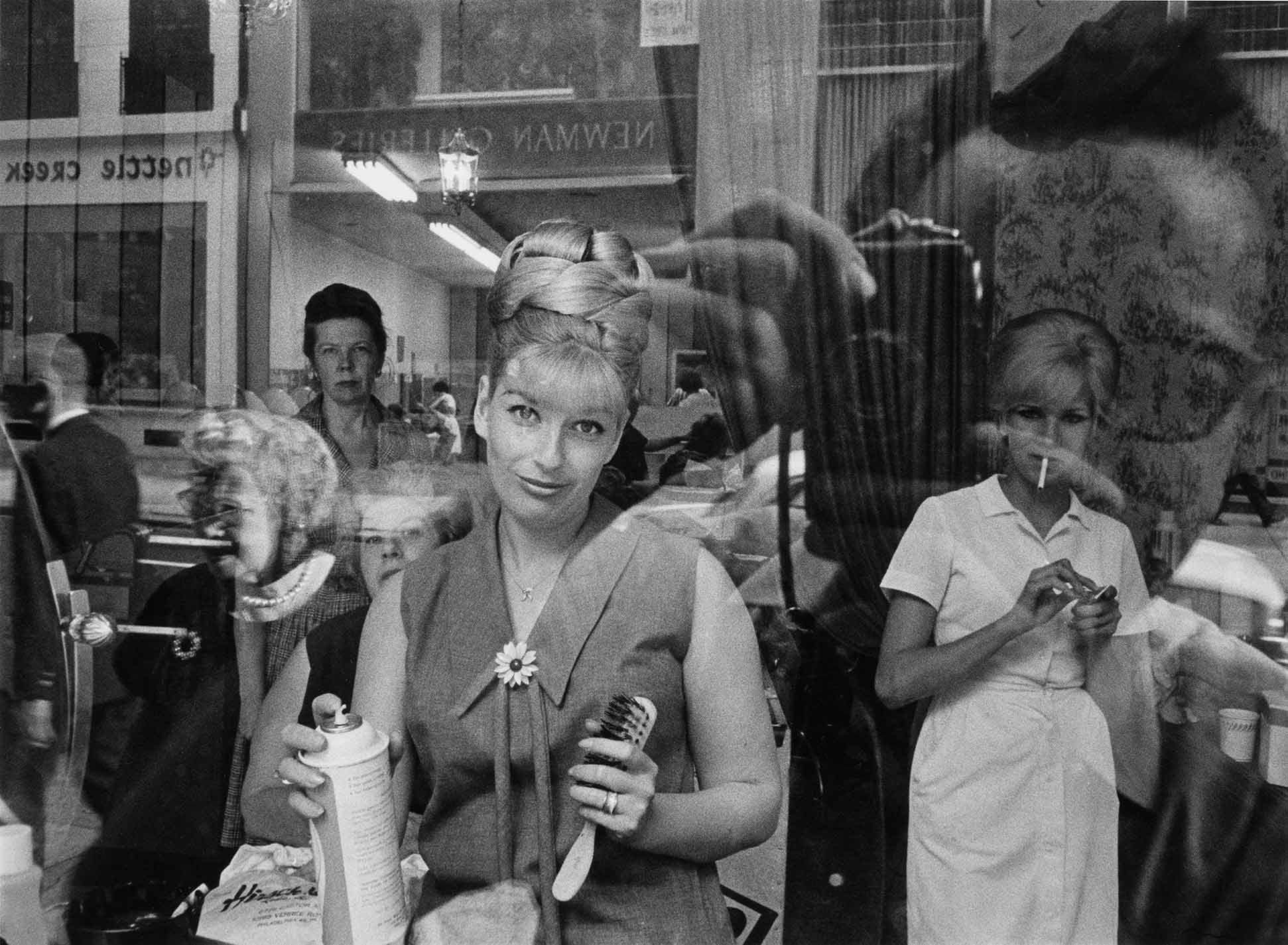 Peering into a beauty shop window, a worker holding a brush and hairspray along with several patrons pause to observe the photographer reflected in the glass.