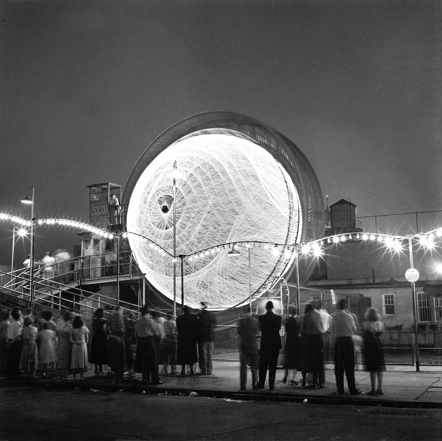 In a long-exposure night scene, a glowing orb appears to spin, creating geometric patterns, as nearby crowds observe from the street beneath twinkling lights.