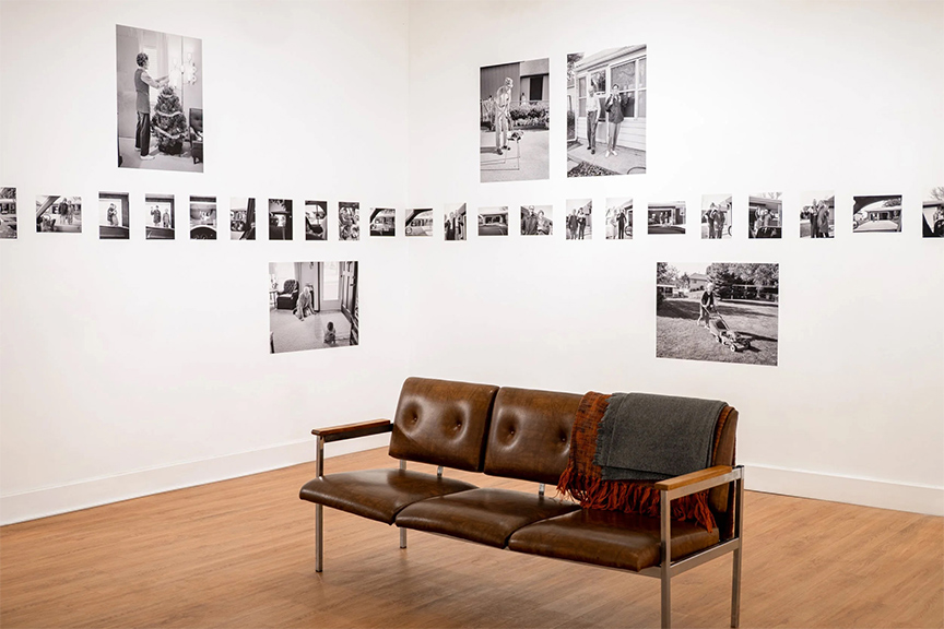 Gallery view of black-and-white photographs arranged in a horizontal line across two white walls, with larger prints spaced above and below, and a brown leather bench with a folded blanket placed in the center of the room.