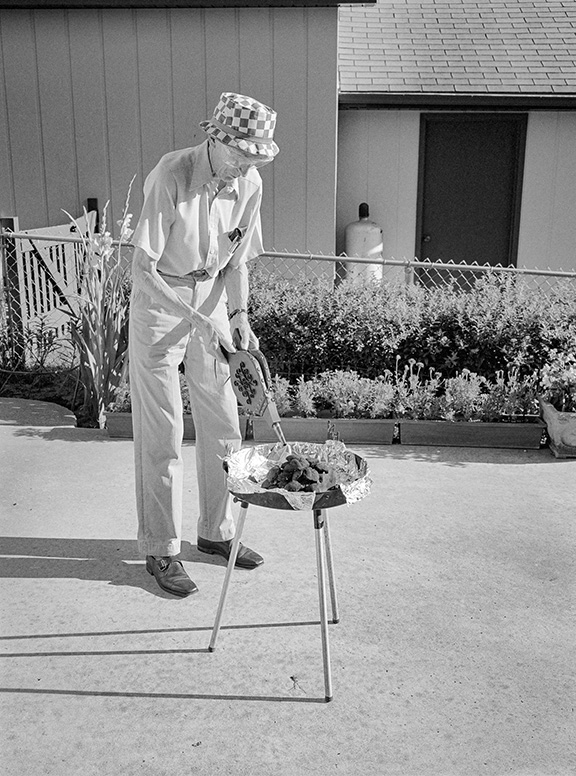 An elderly man wearing a checkered hat uses bellows to tend charcoal in a small grill on a patio, with a garden and house behind him.