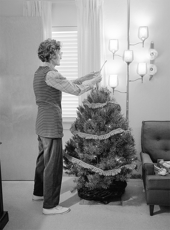 An elderly woman decorates a small Christmas tree in her living room, placing an ornament on top while wall lights glow behind her.