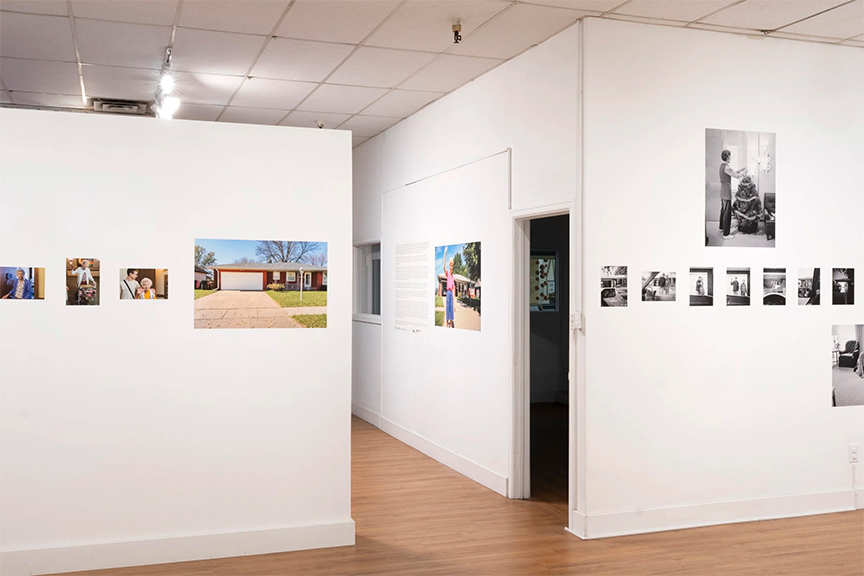 Gallery view of a white-walled exhibition space with photographs arranged in a horizontal line, including color images of a red house and black-and-white portraits, with a doorway opening into an adjacent room.
