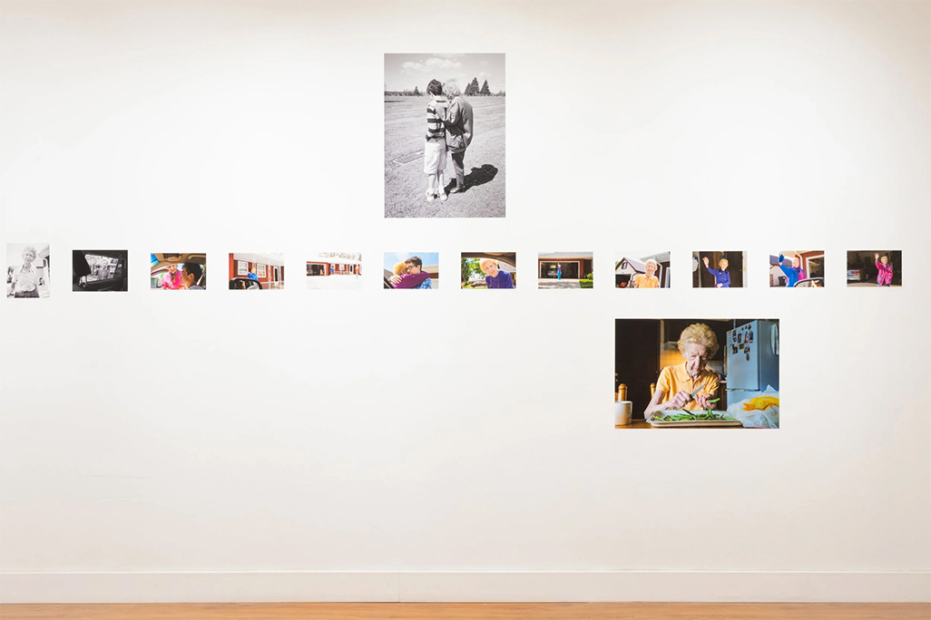 Gallery wall displaying a horizontal row of small color and black-and-white photographs, with a larger black-and-white image centered above and a larger color photograph of an elderly woman seated at a kitchen table mounted below to the right.
