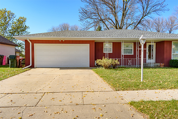 A red single-story house with a white garage door and small front porch sits beneath a clear blue sky, with a concrete driveway and a lamppost on the lawn in front.