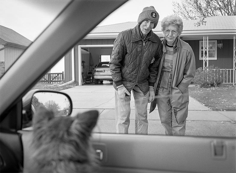 An elderly couple stands arm in arm in their driveway looking toward a dog inside a car, seen from the car’s interior with the house and open garage behind them.