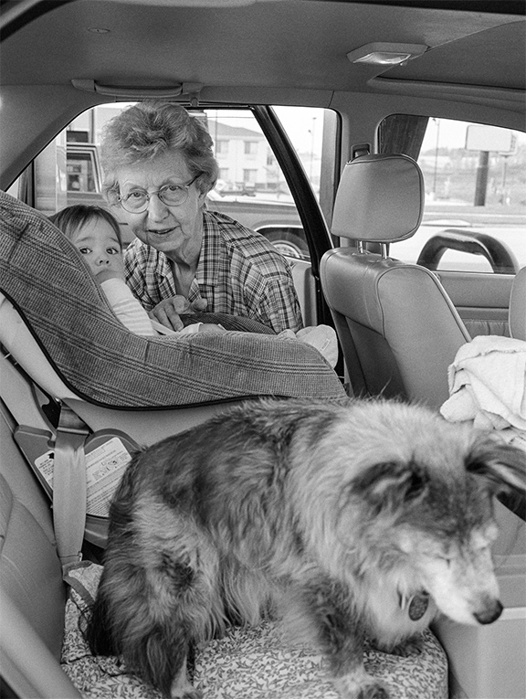 An elderly woman leans into the back seat of a car toward a child in a car seat, while a small dog sits on the front seat in the foreground.