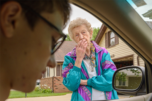 A person wearing glasses sits inside a car looking down as an elderly woman in a teal and purple windbreaker stands outside the window, peering in with her hand over her mouth.