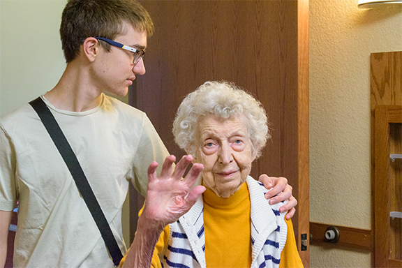 An elderly woman in a yellow sweater and striped vest raises her hand in a small wave while a young man with glasses stands beside her with his arm around her shoulders.