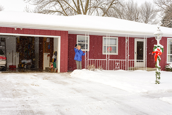 On a snowy day, an elderly woman in a blue coat waves from the porch of a red single-story house decorated for the holidays.