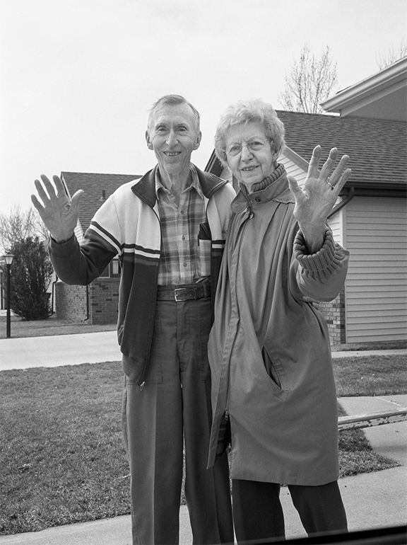An elderly couple stands arm in arm outside their home, smiling and waving toward the viewer.