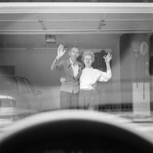 An elderly couple stands arm in arm in their open garage, waving toward the viewer, seen from inside a car with the steering wheel visible in the foreground.