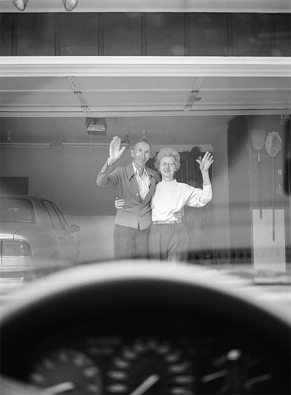 An elderly couple stands arm in arm in their open garage, waving toward the viewer, seen from inside a car with the steering wheel visible in the foreground.