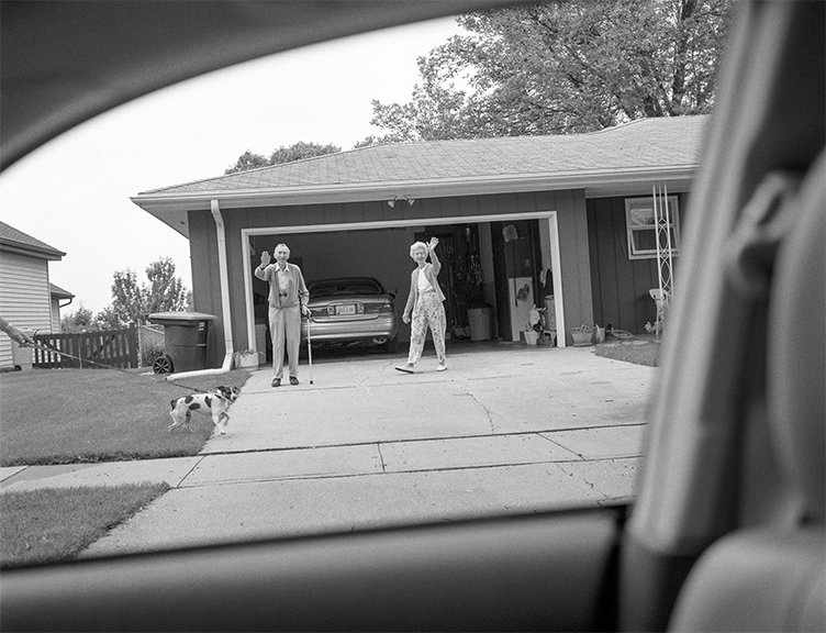 An elderly couple stands in their driveway waving as the viewer observes from inside a car, with a small dog crossing the lawn and an open garage with a parked car behind them.
