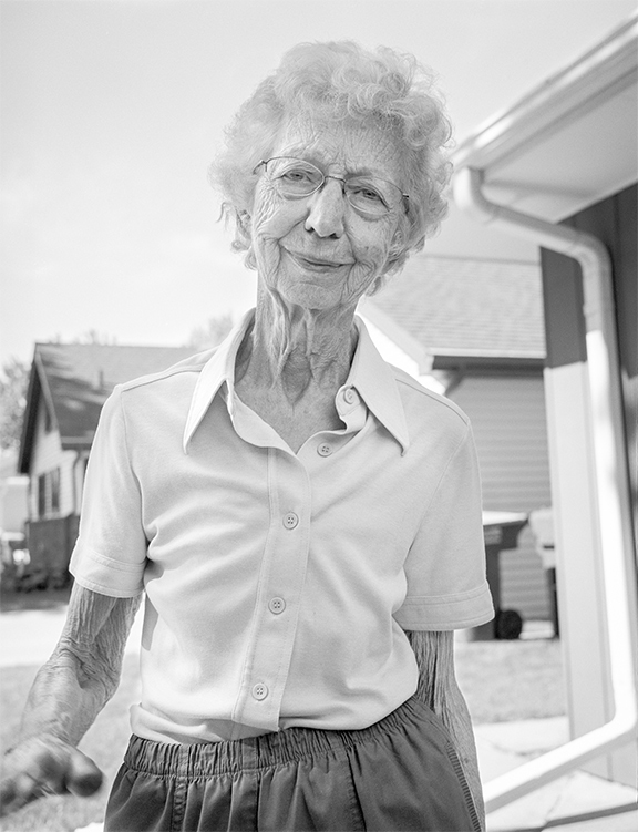 Close-up of an elderly woman wearing a short-sleeve button-up shirt standing outdoors near a garage, looking toward the viewer with a gentle expression.