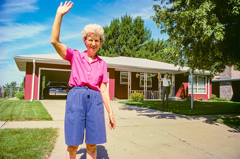 An elderly woman in a pink shirt and blue shorts waves while standing in a driveway, with a man waving from the porch of a red suburban house behind her.