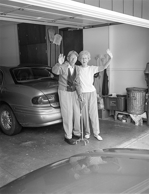 An elderly couple stands together in their garage waving toward the viewer, the man holding a cane, with a parked car and storage bins behind them.