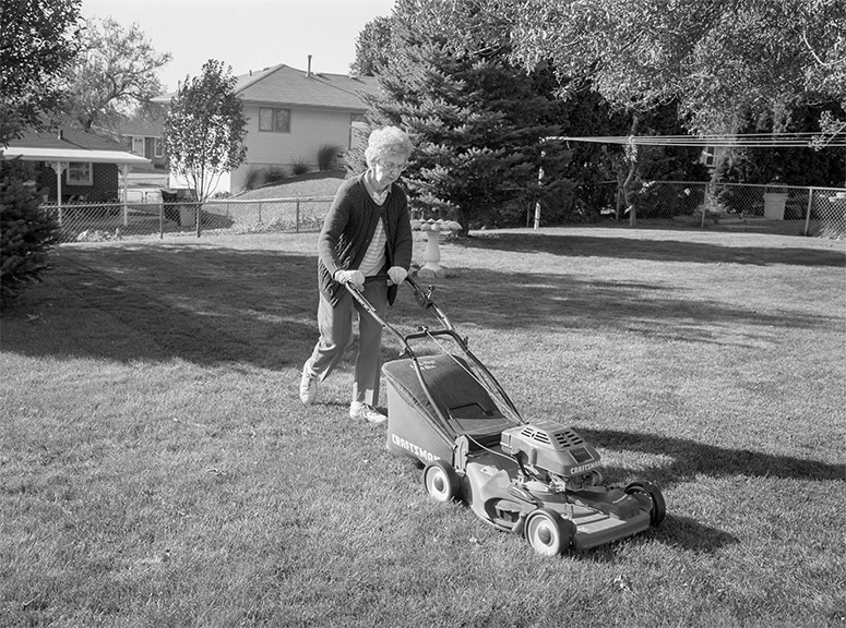 An elderly woman wearing a cardigan and slacks pushes a lawnmower across the yard.